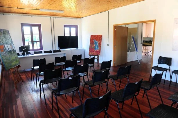 Foto de um auditório contendo várias cadeirass e ao fundo quadros, uma televisão, um computador e uma mesa