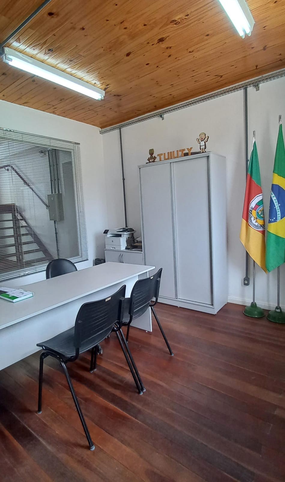 Foto da recepção e gabinete da Subprefeitura de Tuiuty mostrando uma mesa, um armário, bandeiras e uma janela ao fundo.