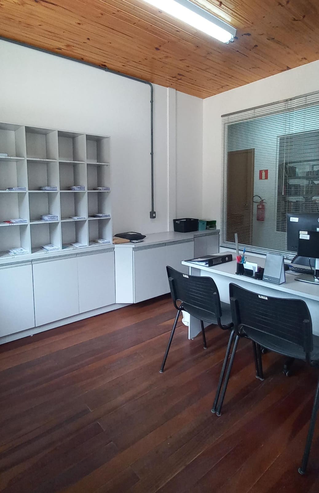 Foto da recepção e gabinete da Subprefeitura de Tuiuty mostrando uma mesa, uma estante e uma janela ao fundo.