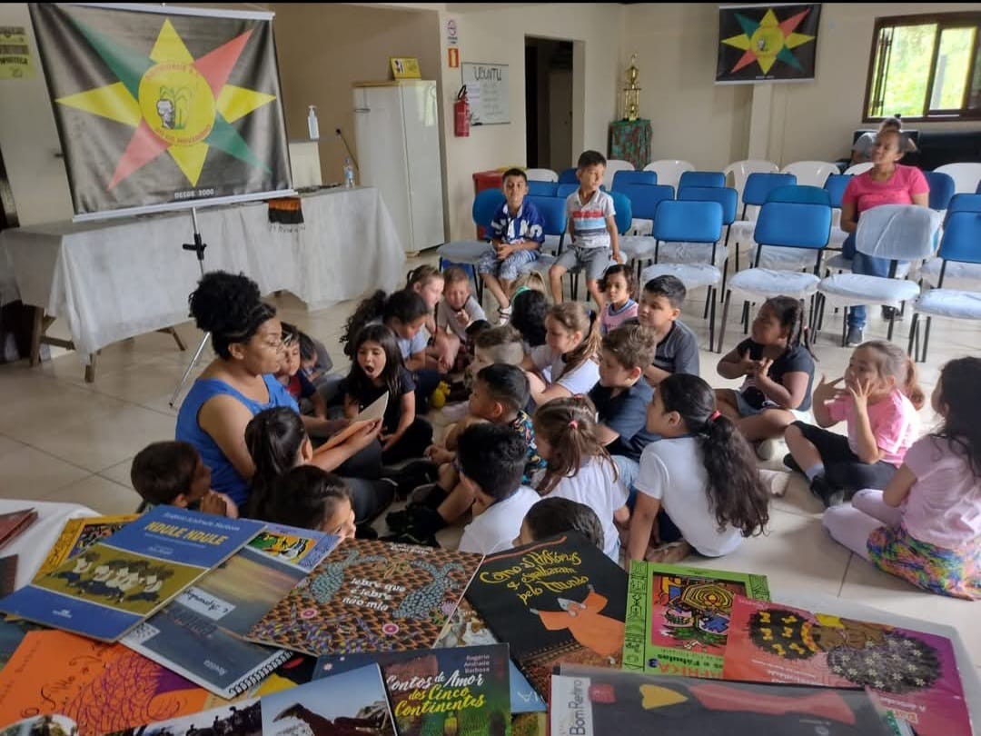 Foto da sala de convivência contendo cadeiras ao fundo e, em frente delas, diversas crianças sentadas no chão junto de uma professora. Mais à frente, uma mesa cheia de livros