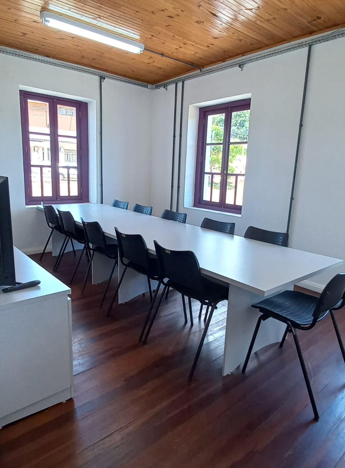 Foto mostrando uma mesa comprida e diversas cadeiras ao seu redor. Duas janelas ao fundo.