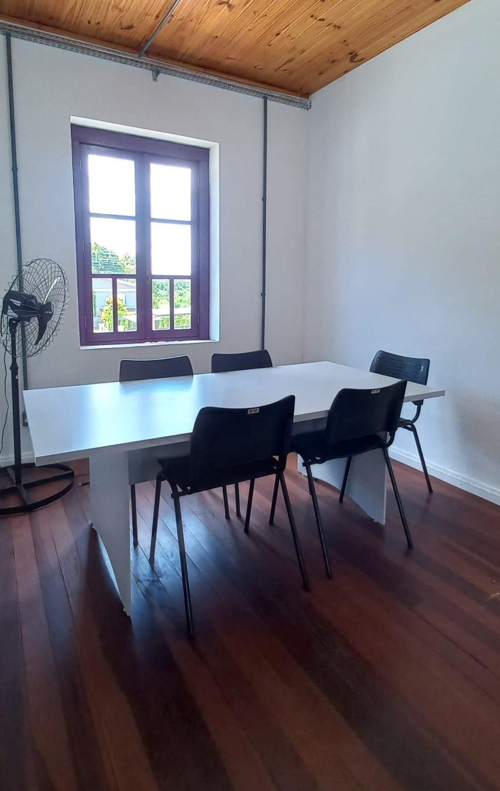 Foto de uma sala com uma mesa e cadeiras em sua volta. Ao fundo, uma janela